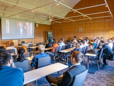 PhD candidates and professors sit in a lecture room and look at a screen showing Professor Tolan, President of the University of Göttingen.