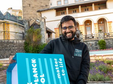 A student holds a seat cube on which the Matter to Life logo is displayed in large letters. Ringberg Castle can be seen in the background.