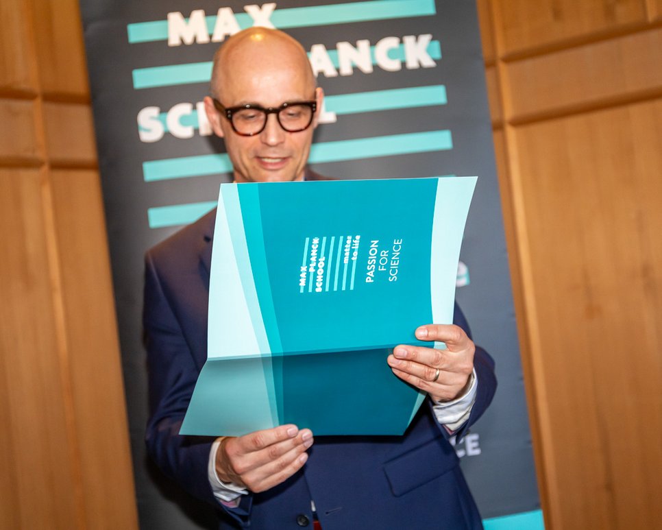 Close up of Prof. Joachim Spatz holds a certificate folder in his hand and reads from it. In the background you can see a roll-up with the Matter to Life logo