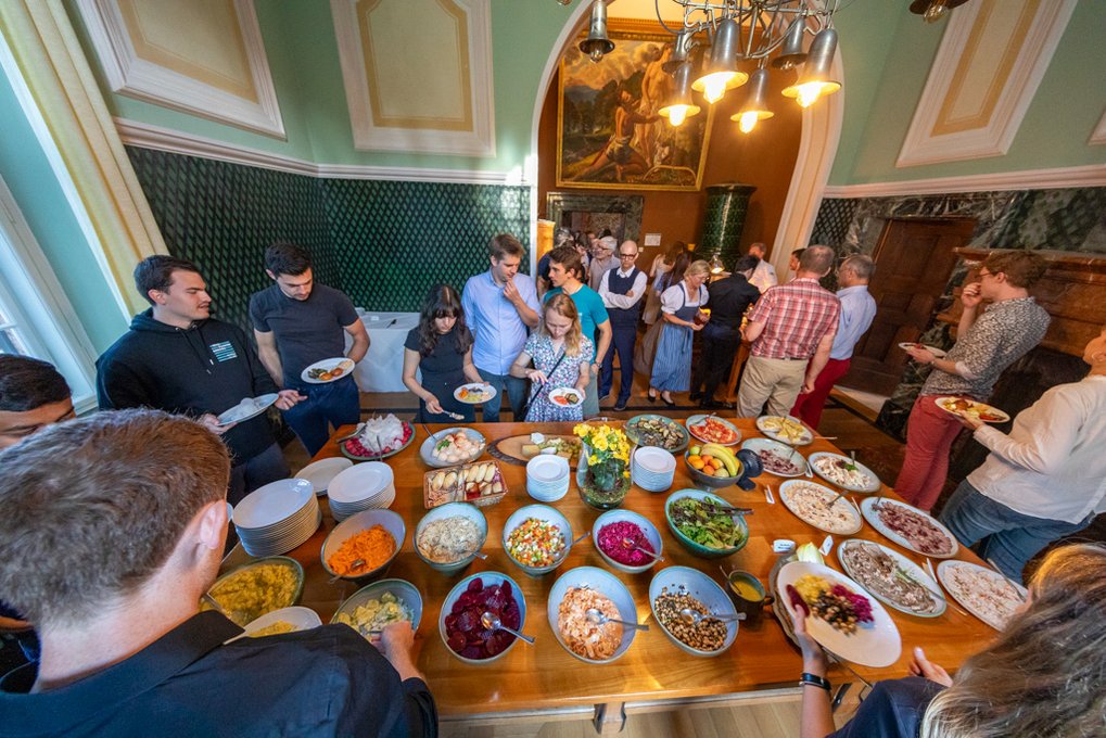 You can see a table with many different colorful salads. Surrounded by lots of people getting something to eat. 