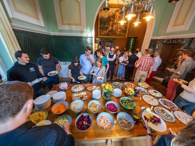 You can see a table with many different colorful salads. Surrounded by lots of people getting something to eat.