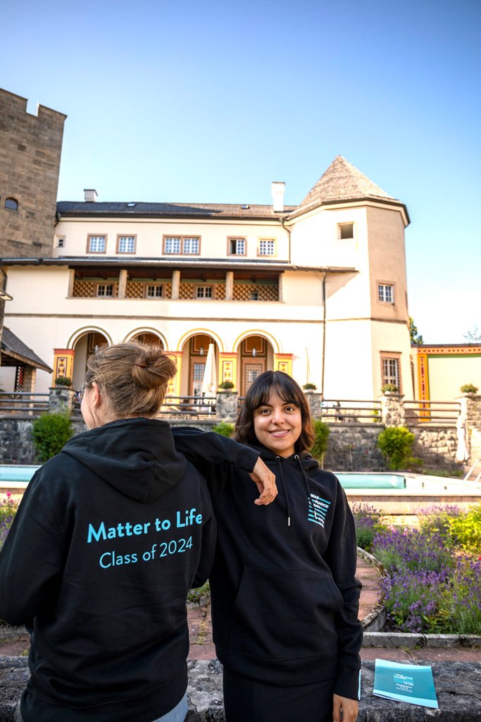 You can see two PhD candidates, one from behind and the other from the front. The latter puts her elbow on the other's shoulder and smiles at the camera. Both are wearing a black Matter to Life hoodie. The words “Matter to Life Class of 2024” can be seen on the back of the hoodie. Ringberg Castle can be seen in the background. 