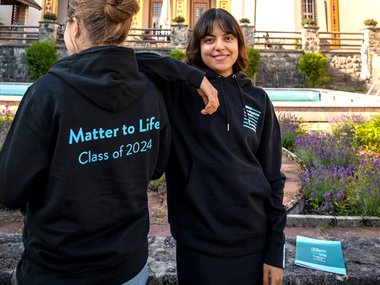 You can see two PhD candidates, one from behind and the other from the front. The latter puts her elbow on the other's shoulder and smiles at the camera. Both are wearing a black Matter to Life hoodie. The words “Matter to Life Class of 2024” can be seen on the back of the hoodie. Ringberg Castle can be seen in the background.