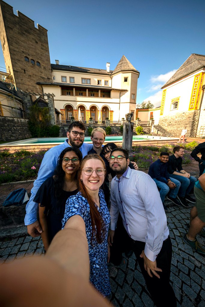 Three female and three male students take a selfie. The scenery of Ringberg Castle can be seen in the background
