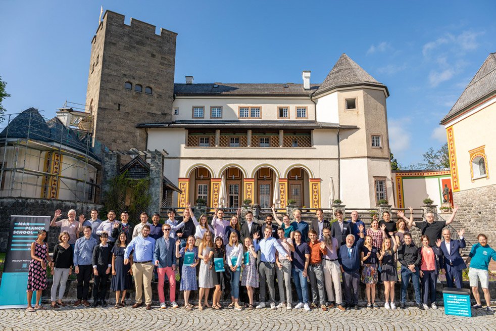 This is a group photo of 45 participants of the Ringberg Symposium, with Ringberg Castle in the background. The people in the photo are posing happily. 