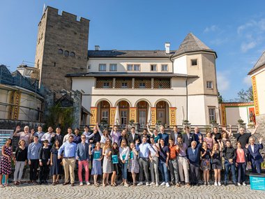 This is a group photo of 45 participants of the Ringberg Symposium, with Ringberg Castle in the background. The people in the photo are posing happily.
