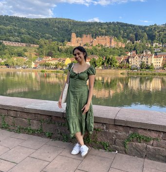 Lasya Damaraju is posing and smiling in front of the camera in front of a river and a castle nestled in a green landscape.