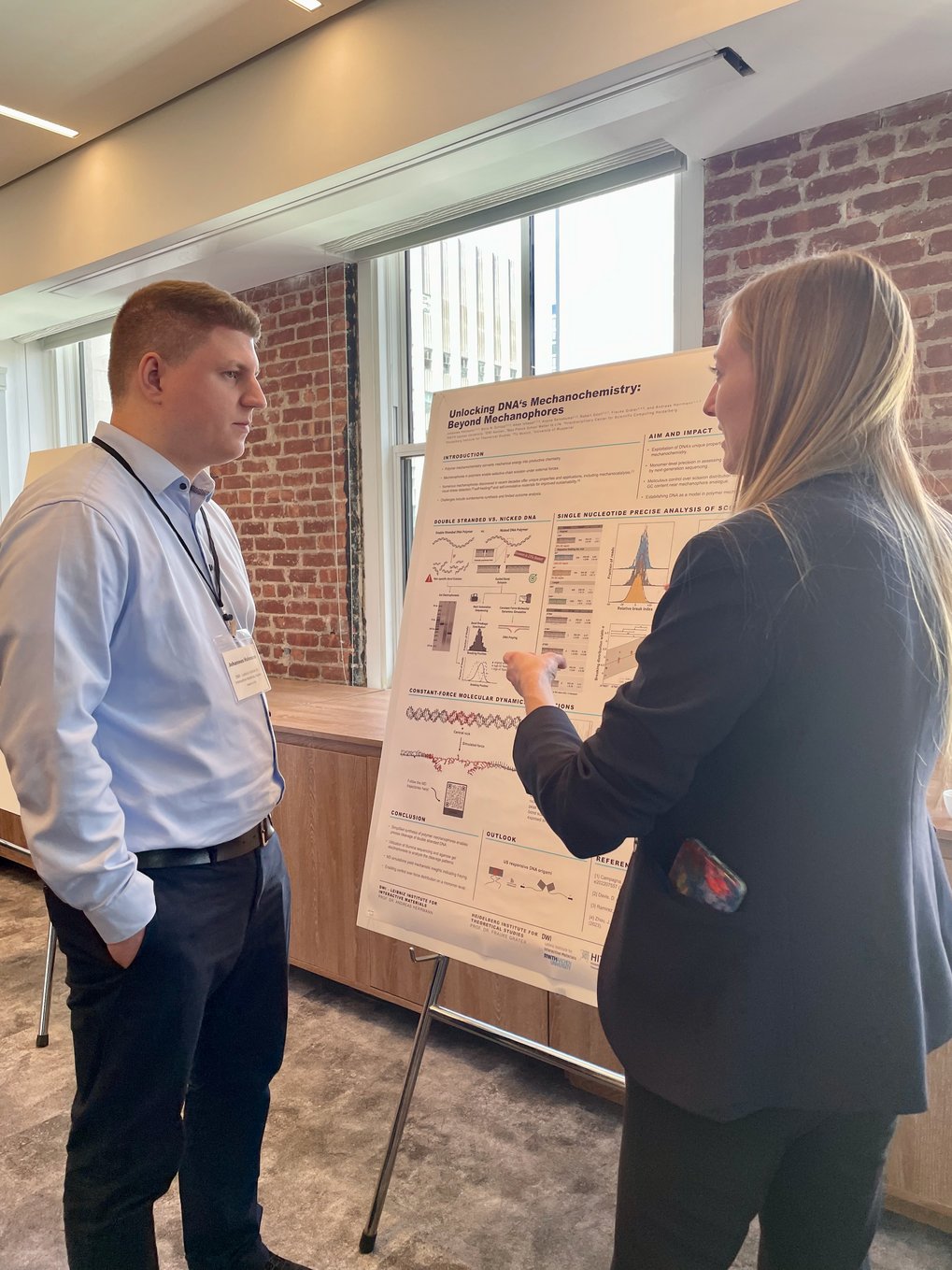 Two people are standing in front of a poster and discussing its content in a room with brick walls and large windows.