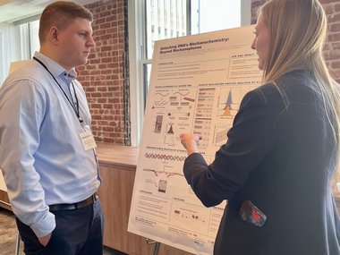Two people are standing in front of a poster and discussing its content in a room with brick walls and large windows.