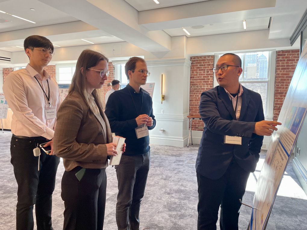 A person presents information on a board to an attentive group of three people in a light-flooded room. 