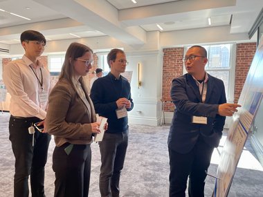 A person presents information on a board to an attentive group of three people in a light-flooded room.