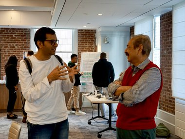 Two men discussing in a conference room, with a table and chairs visible in the background.