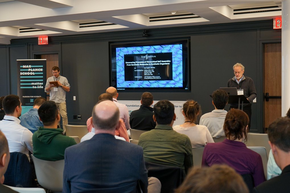A conference room with a speaker presenting at a podium and a large screen, with attendees seated and a 'Matter to Life' banner in the background.