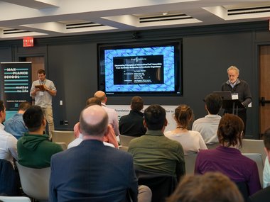 A conference room with a speaker presenting at a podium and a large screen, with attendees seated and a 'Matter to Life' banner in the background.