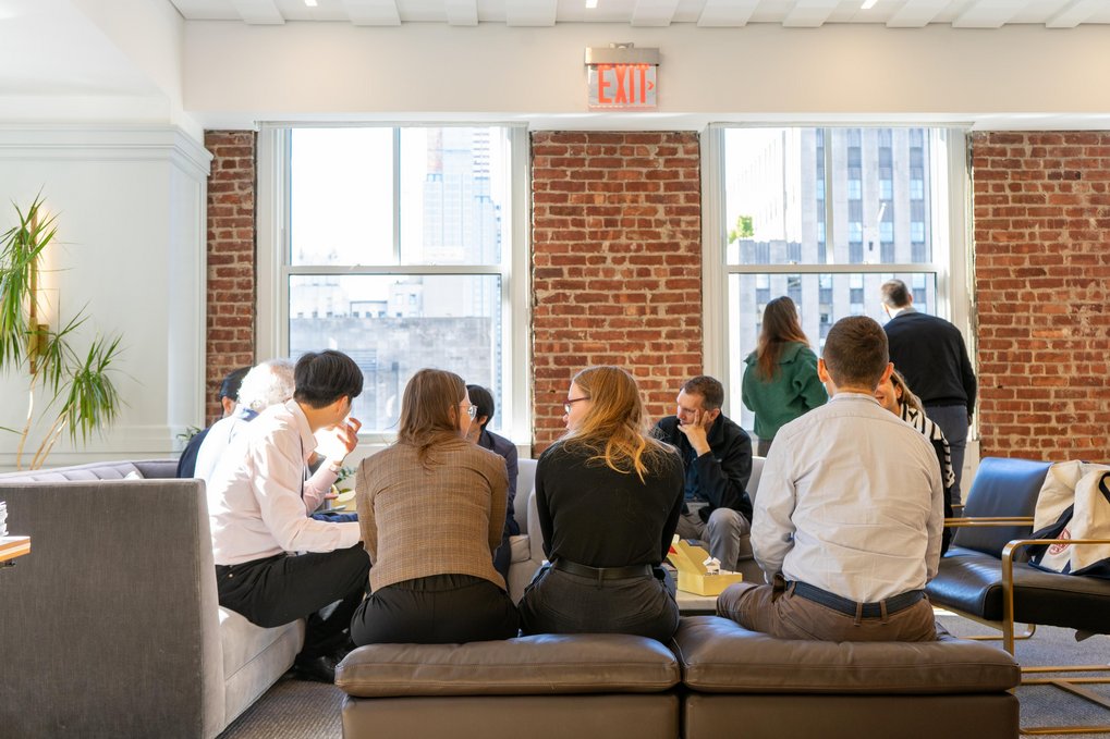 People seated on sofas and chairs in a bright room with brick walls and large windows, engaged in small group discussions, while a few individuals stand near the windows
