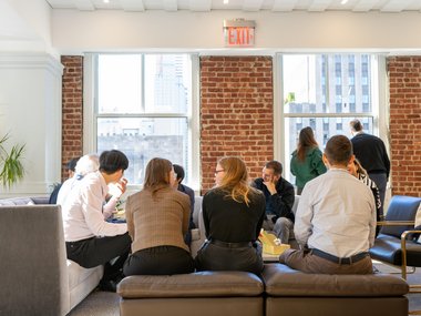 People seated on sofas and chairs in a bright room with brick walls and large windows, engaged in small group discussions, while a few individuals stand near the windows