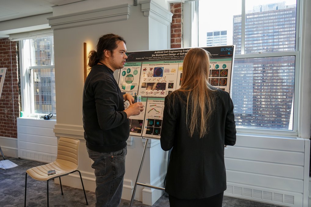 Two people are standing in front of a poster and discussing its content in a room with brick walls and large windows overlooking the city.