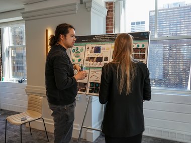 Two people are standing in front of a poster and discussing its content in a room with brick walls and large windows overlooking the city.