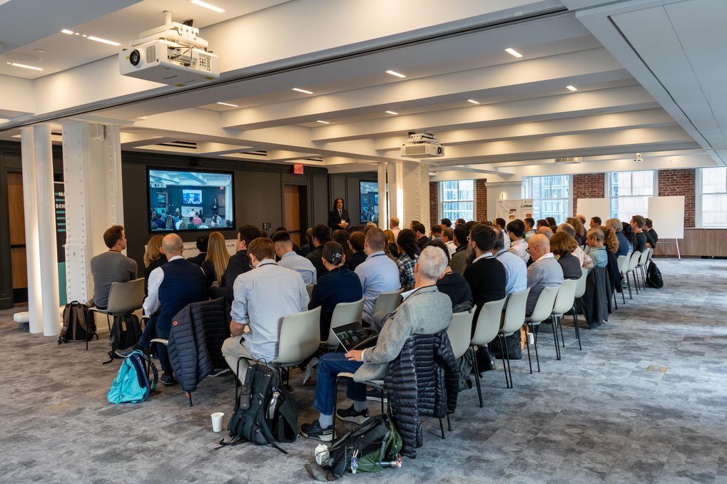 A large group of the Matter-to-Life conference participants seated in rows in a modern conference room, watching a presentation displayed on a screen at the front of the room