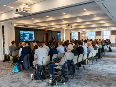 A large group of the Matter-to-Life conference participants seated in rows in a modern conference room, watching a presentation displayed on a screen at the front of the room