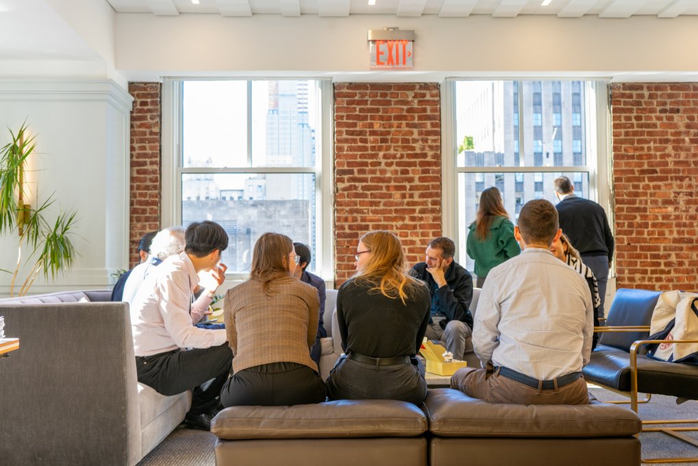 Several people are seated in a modern office lounge with brick walls, discussing work. Sunlight streams through the large windows, illuminating the room.