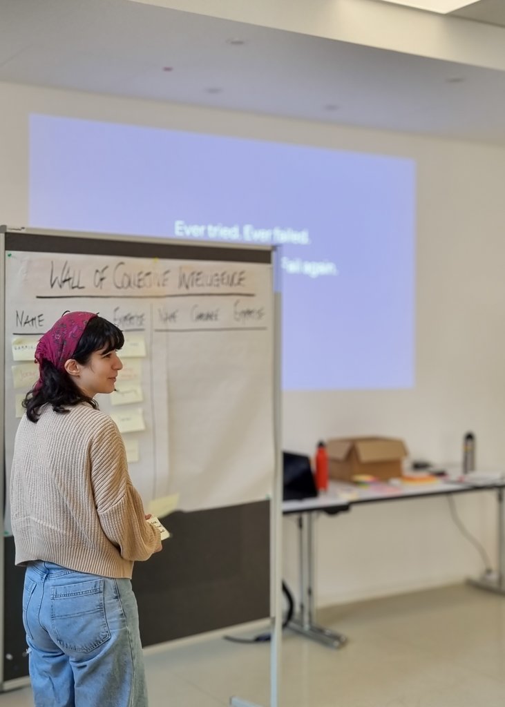Participant standing near a board titled 'Wall of Collective Intelligence,' holding sticky notes, with a projected motivational quote in the background