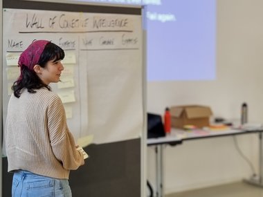 Participant standing near a board titled 'Wall of Collective Intelligence,' holding sticky notes, with a projected motivational quote in the background