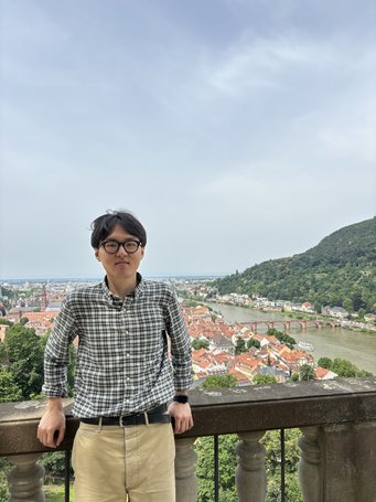 Won Joon standing on a viewing terrace overlooking the picturesque city of Heidelberg, Germany with its distinctive red rooftops, lush green trees, and the Neckar River.