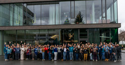 A diverse group of people, numbering around 100, stand together in front of a building with large glass windows, labeled "Campus Garden," many waving at the camera.