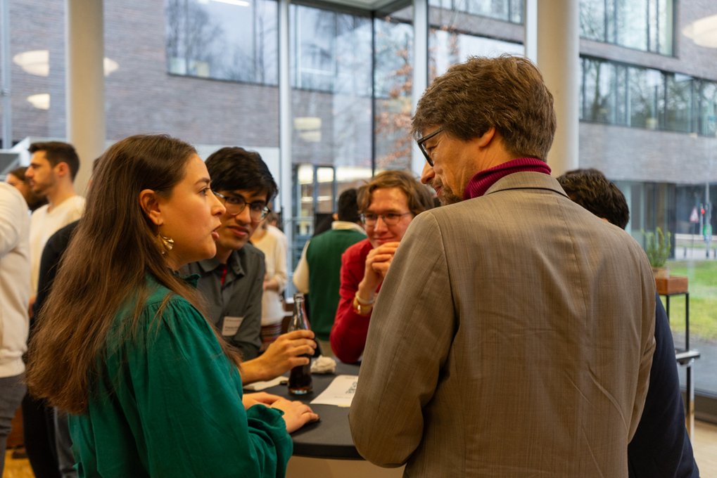 People discussing, some holding drinks, against a backdrop of large windows and modern architecture.