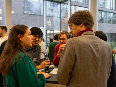 People discussing, some holding drinks, against a backdrop of large windows and modern architecture.