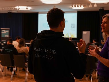 In a conference room, participants are seated facing a presentation screen. A person wearing a "Matter to Life Class of 2024" hoodie engages in conversation.
