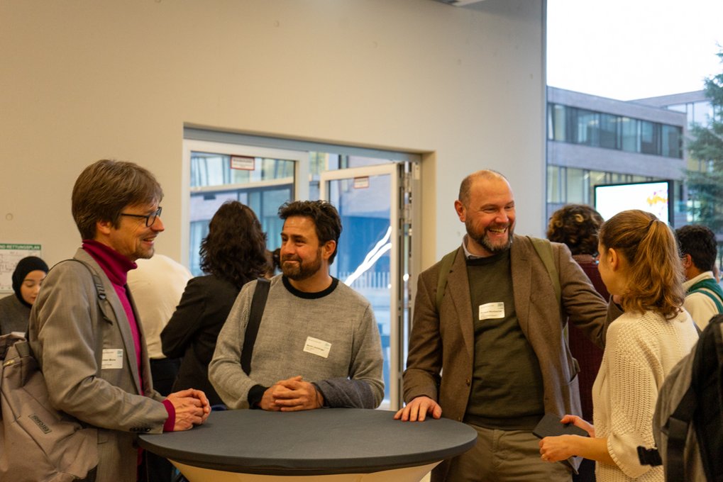 Several individuals are conversing around a standing table. They wear casual attire with visible name tags. The setting includes an open door, leading to a modern building exterior.
