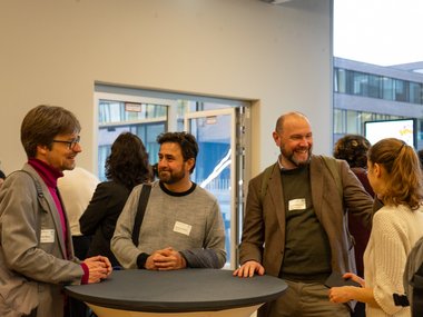 Several individuals are conversing around a standing table. They wear casual attire with visible name tags. The setting includes an open door, leading to a modern building exterior.