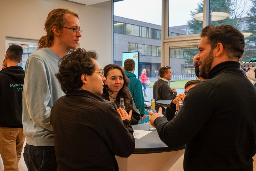  Several people are gathered around a high table indoors, engaged in discussion. Bottles are on the table, and large windows provide a view of the outside, where a modern building and people walking can be seen.