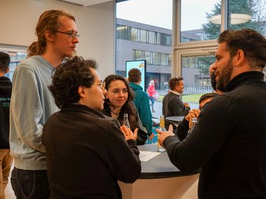 Several people are gathered around a high table indoors, engaged in discussion. Bottles are on the table, and large windows provide a view of the outside, where a modern building and people walking can be seen.