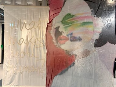 Installation with two fabric panels: front displays a blurred face painted in rainbow hues, rear panel features embroidered philosophical question "What makes you human?" in elegant cursive.
