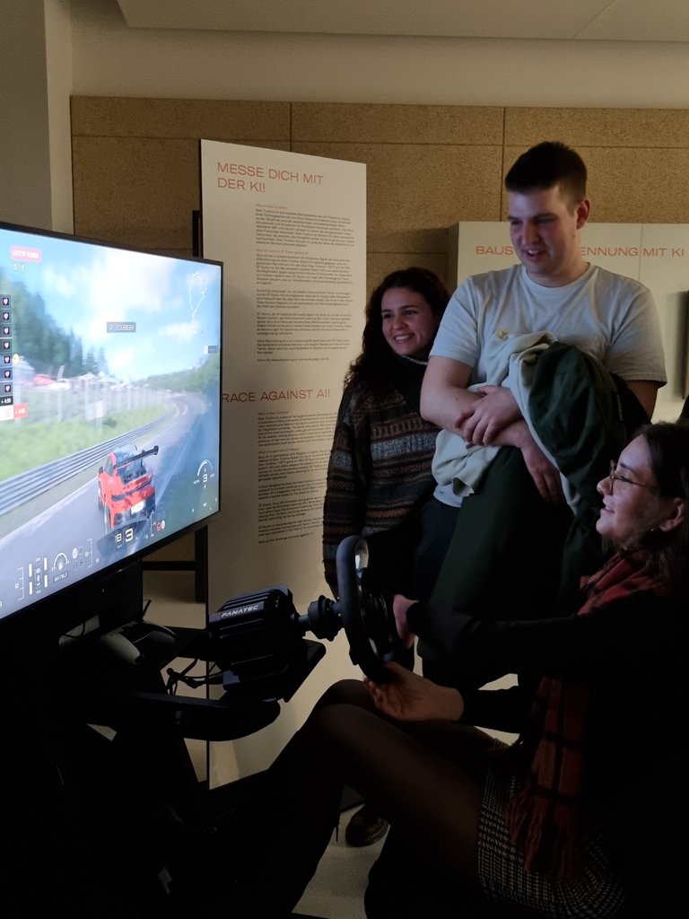 A woman sits at a racing simulator setup, holding a steering wheel and focused on the driving game displayed on a large screen. Two young adults, a man and a woman, stand nearby smiling and watching. The scene takes place in an indoor exhibition or event space with informational posters on the wall in the background.