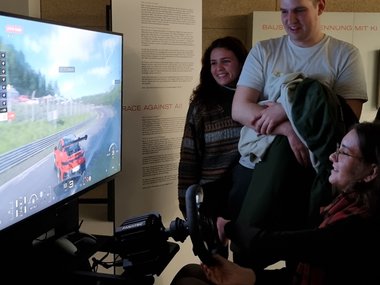 A woman sits at a racing simulator setup, holding a steering wheel and focused on the driving game displayed on a large screen. Two young adults, a man and a woman, stand nearby smiling and watching. The scene takes place in an indoor exhibition or event space with informational posters on the wall in the background.