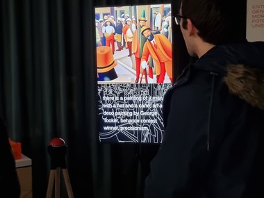A person in a dark coat looks at a work of art on a large screen. The painting shows a group of people in vivid colors and geometric shapes. Curtains can be seen in the background.