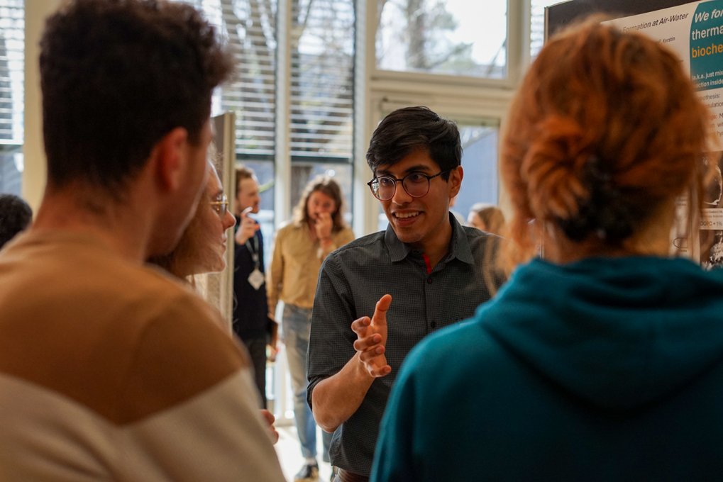 Four individuals engaged in conversation near a scientific poster in a sunlit space.