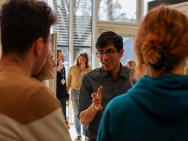 Four individuals engaged in conversation near a scientific poster in a sunlit space.