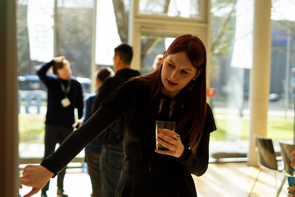 A Matter to Life PhD candidate, dressed in black, gestures towards a display or board while holding a drink, possibly explaining something. She is indoors in a bright, modern space with large windows, and several people are conversing in the background.