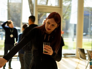 A Matter to Life PhD candidate, dressed in black, gestures towards a display or board while holding a drink, possibly explaining something. She is indoors in a bright, modern space with large windows, and several people are conversing in the background.