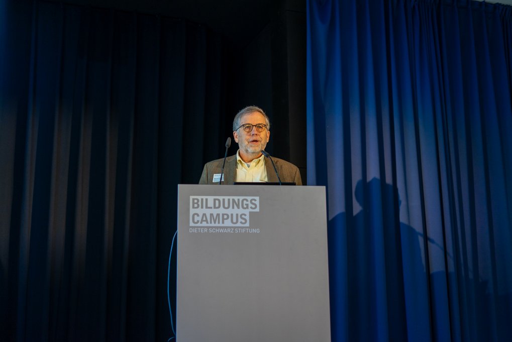 Michael Donihue at podium with "Bildungscampus" logo, dark curtains in background.