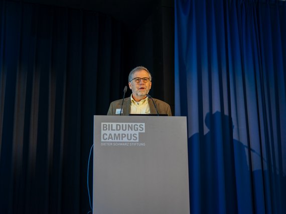 Michael Donihue at podium with "Bildungscampus" logo, dark curtains in background.