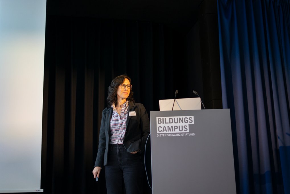 Roseanne Zia at podium with "Bildungscampus" logo, dark curtains in background.