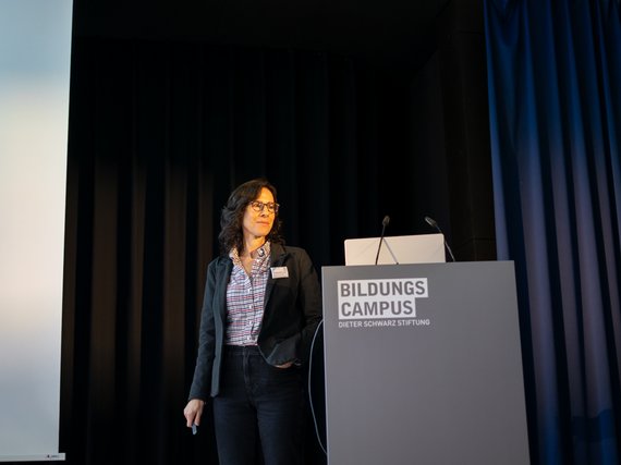 Roseanne Zia at podium with "Bildungscampus" logo, dark curtains in background.