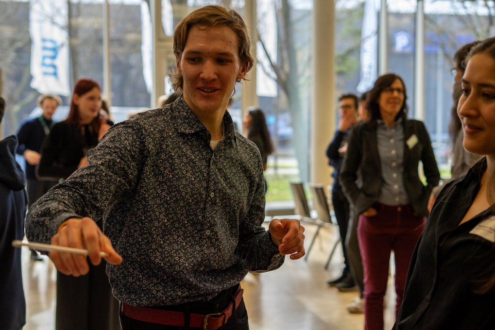 Matter to Life PhD candidates in business attire interacting at a professional gathering, with a focus on one person gesturing with a pen.