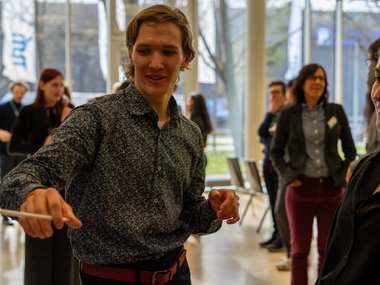 Matter to Life PhD candidates in business attire interacting at a professional gathering, with a focus on one person gesturing with a pen.